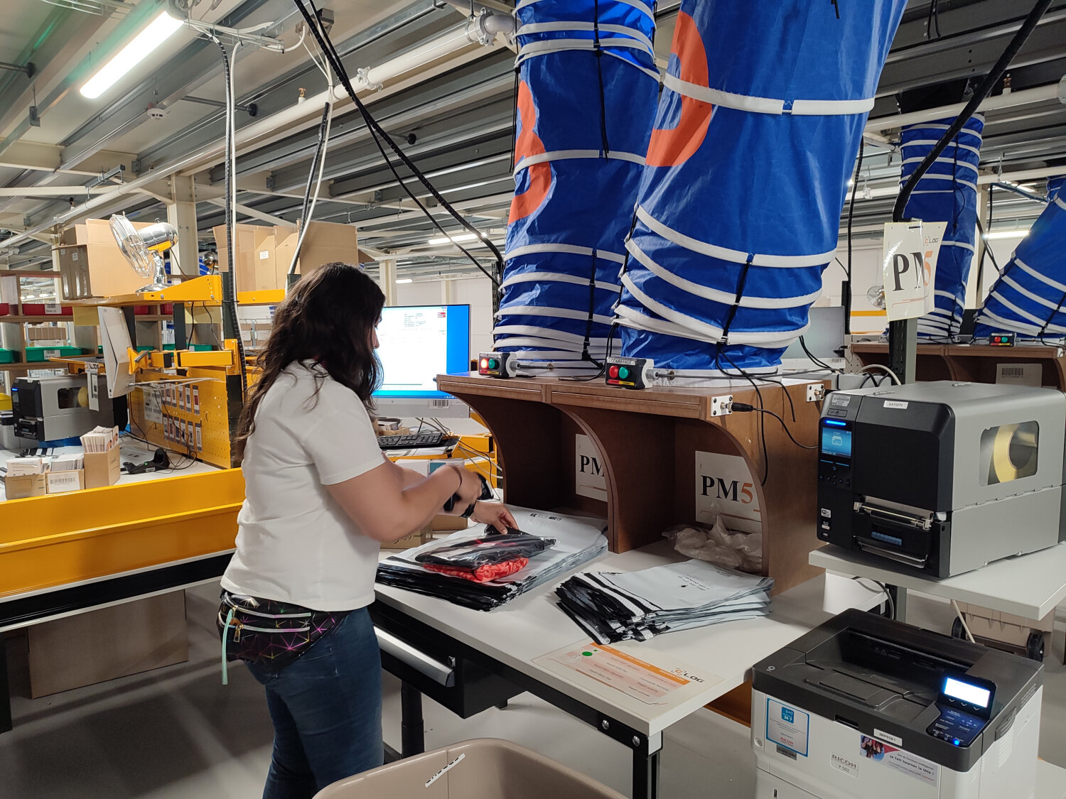 Les robots viennent faciliter le travail des équipes. Ici, l’opératrice s’occupe d’emballer plusieurs articles d’une même commande déposés par les robots via le toboggan bleu. - © Républik Retail