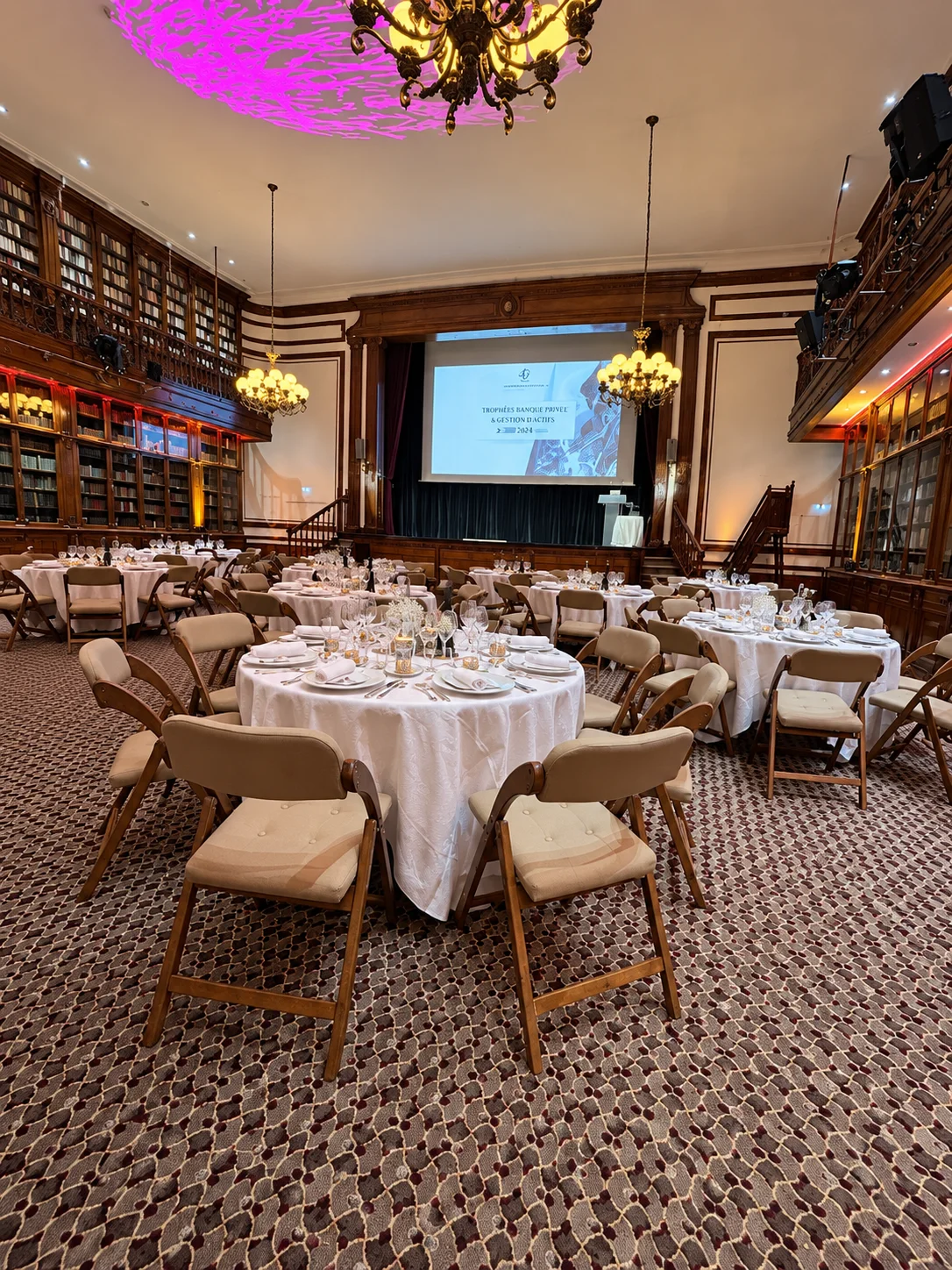 La salle de bibliothèque dressée pour le dîner : tables rondes, cristal et miroirs, sous les boiseries et les lustres.